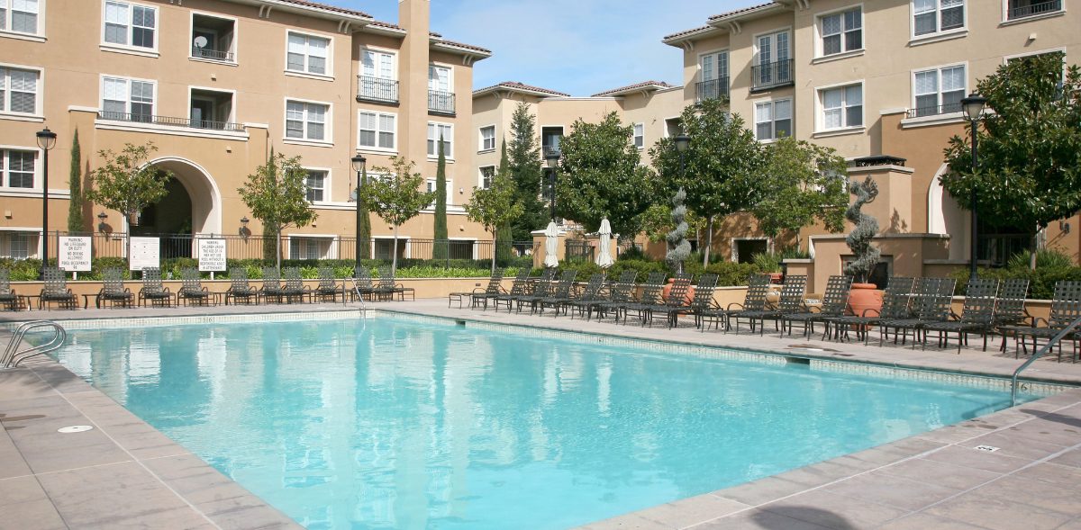 Photo of a modern apartment complex with beige exterior, multiple balconies, and landscaped trees. In the foreground is a large outdoor swimming pool surrounded by lounge chairs and umbrellas, creating a resort-style amenity for residents